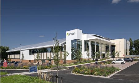 Modern leisure centre building with glass facade and landscaped surroundings, featuring parking spaces and greenery in the foreground.