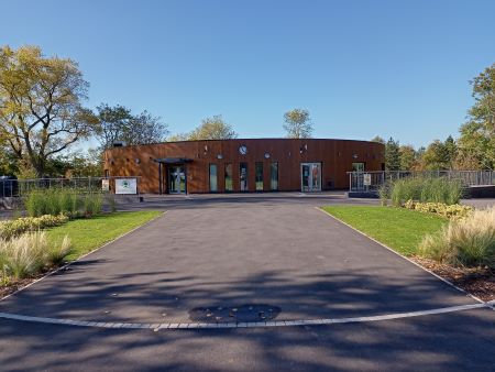 Tarmac driveway with greenery either side, leading up to a circular community pavilion clad in wood.