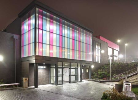 Wednesbury Leisure Centre exterior at night, featuring colorful illuminated glass panels and entrance doors, with surrounding pathways and landscaping.